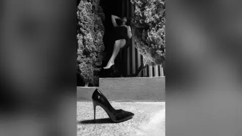 Elegant woman in a black dress posing by lush trees in a moody black and white outdoor setting.