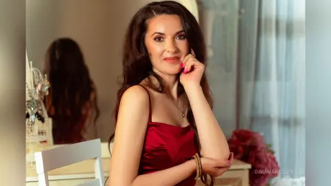 DaianaKelley is a brunette woman wearing a red satin camisole, posing with her hand on her cheek in a bright room.