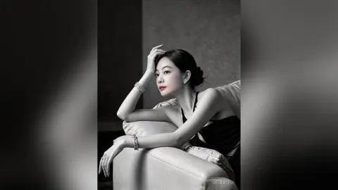 CelesteQi — CelesteQi poses in a black dress while leaning against a light-colored armchair.