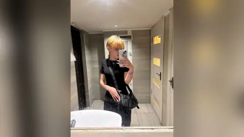 Blonde woman in a black t-shirt taking a mirror selfie in a modern bathroom setting.