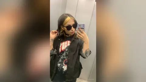 Trendy woman with blonde highlights in a graphic tee and sunglasses posing for a mirror selfie in an elevator.