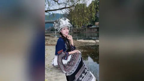 Young woman in traditional silver ethnic headpiece and patterned blue dress by a serene river landscape.