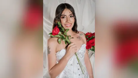 Smiling woman in white lace corset and gold headpiece holding red roses in a soft, romantic studio setting.
