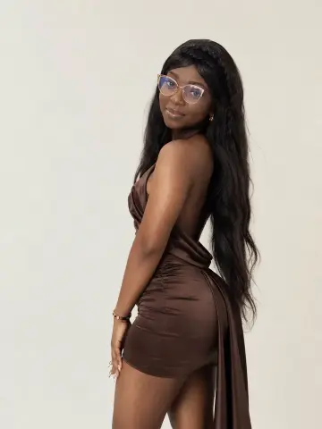 Smiling Black woman in a brown satin cocktail dress and glasses posing against a white studio background.