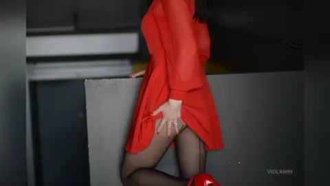 ViolaWin — ViolaWin wears a short red dress and black tights while posing against a dark wall.