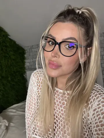 Blonde woman with glasses wearing a white mesh top in a cozy room with a green moss wall.