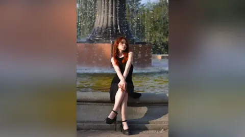Redheaded woman in a black sleeveless dress sitting by a stone fountain in a serene outdoor park setting.