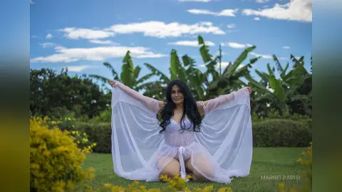 Smiling woman in a flowing white ethereal gown sitting in a lush tropical garden under a blue sky.