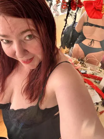 Smiling redhead woman in black lace lingerie relaxing in a sunlit bedroom setting.