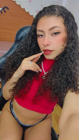 Young woman with long curly hair wearing a red top and silver necklace in a cozy indoor setting.