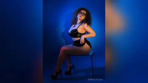 Curvy woman in black lingerie sitting on a chair against a moody blue studio background.