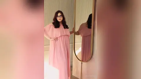 Elegant woman in a flowing pink maxi dress posing in a bright, minimalist room with a gold mirror.