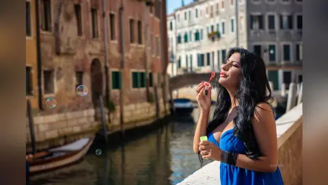 MissyJolie poses outdoors by a Venice canal, blowing bubbles while wearing a blue sundress.