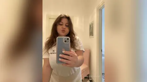 Young woman in a white graphic t-shirt taking a casual selfie in a bright, minimalist bedroom.