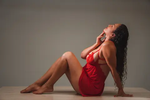 Curvy woman in a red lace bodysuit sitting on a floor against a neutral studio background with a moody vibe.
