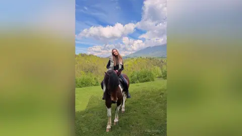 EvelynMills sits upright on a brown horse in a grassy field under a blue cloudy sky.