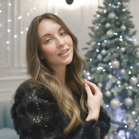 Smiling young woman in a black sparkly sweater posing in front of a glowing Christmas tree.