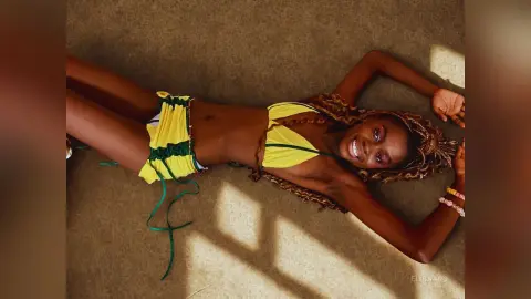 Smiling Black woman in a yellow bikini relaxing on a sunlit carpeted floor.