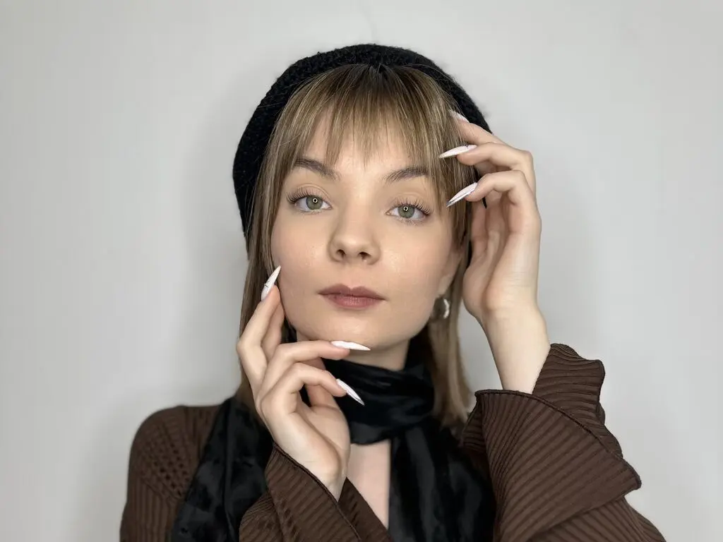 Young woman with blonde bangs wearing a black beanie and brown ribbed sweater in a minimalist studio setting.