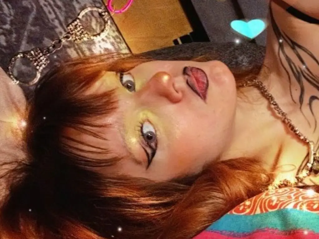 Redheaded woman with dramatic eye makeup and tattoos lying down in warm, moody lighting.