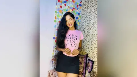 Smiling young woman in a pink graphic t-shirt and black skirt posing against a colorful confetti background.