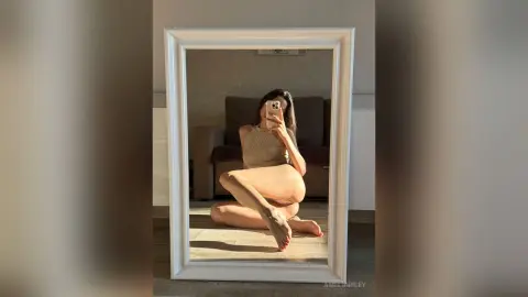 AmeliaRiley poses for a mirror selfie, sitting on the floor in a tan tank top and showing her bare legs and feet.