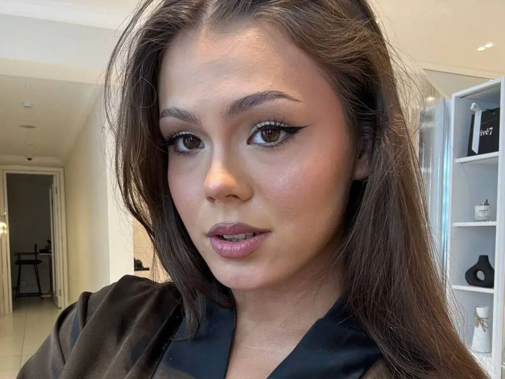 DemetriceTaneja looks directly at the camera with long brown hair and winged eyeliner, wearing a black silk top.