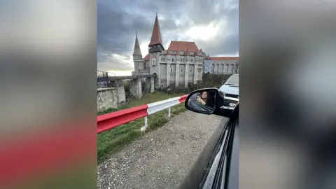 AnaMathers poses in a car side mirror reflection with a large stone castle in the background under a cloudy sky.