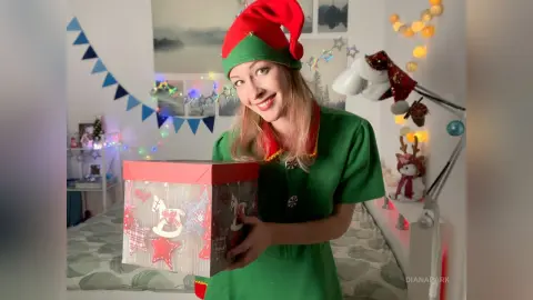 Smiling woman in a green elf costume holding a large red gift box in a festive holiday room setting.