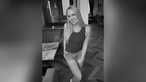 Smiling blonde woman in a dark tank top and denim shorts standing in a modern cafe with herringbone floors.