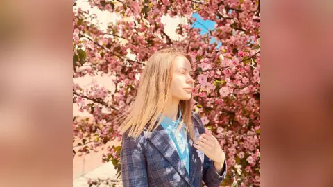 Blonde woman in a blue plaid blazer smelling pink cherry blossoms in a bright spring garden.