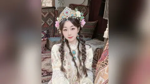 Young woman in traditional ornate white ethnic dress and floral crown sitting in a cozy bohemian room.