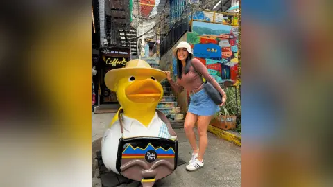 Smiling woman in a brown top and denim skirt posing with a large Pato Pati duck statue on a colorful city street.