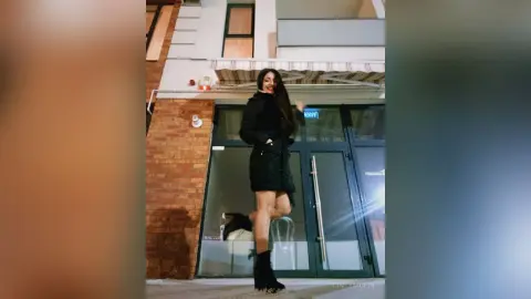Smiling woman in a black winter coat and boots standing outside a modern building at night.