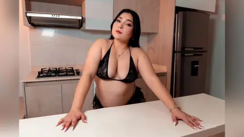 Young woman in black lace bikini lingerie posing confidently in a modern kitchen setting.