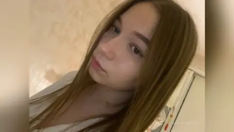 MagalyBoffa looks into the camera with long brown hair and a neutral expression, wearing a white top against a light-colored wall.