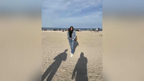 Young woman in a denim jacket and jeans standing on a sunny beach under a blue sky.