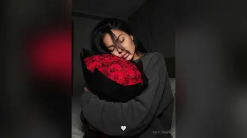 Young woman in a grey sweater hugging a large bouquet of red roses in a moody, dimly lit room.