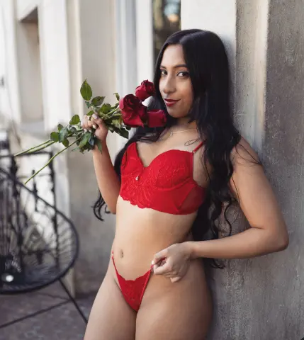 Young woman in red lace lingerie holding red roses while leaning against a stone wall in soft daylight.