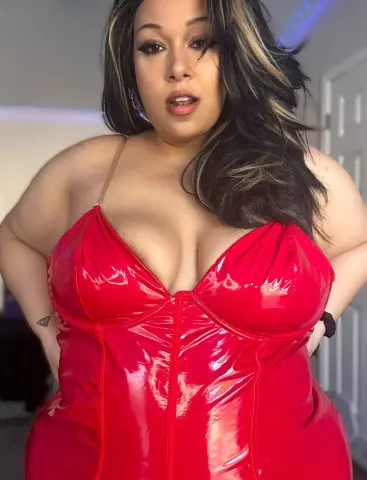 Curvy woman in a red latex corset dress posing confidently in a modern bedroom setting.