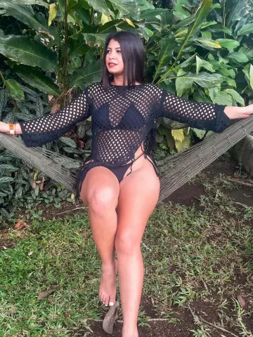 Woman in black mesh cover-up sitting on a hammock in a lush tropical garden setting.