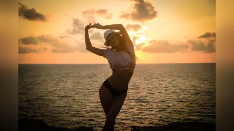 AnaMathers poses against a sunset over the ocean, wearing a white crop top and black bikini bottoms with her arms raised above her head.