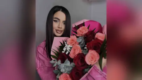 Smiling woman in a pink outfit holding a large bouquet of red and pink roses in a soft, romantic setting.