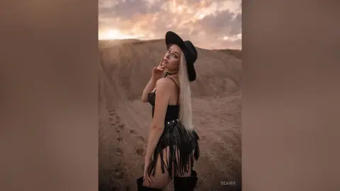 BeaNix poses in a black fringe bodysuit and black cowboy hat against a desert landscape at sunset.