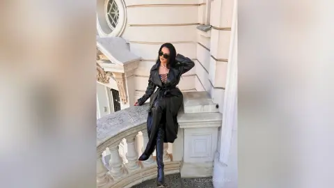 Stylish woman in a black leather trench coat and boots posing on a white stone balcony in bright daylight.