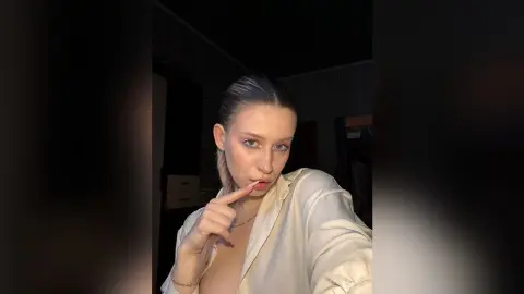 Young woman in a white silk blouse posing with a thoughtful expression in dim indoor lighting.