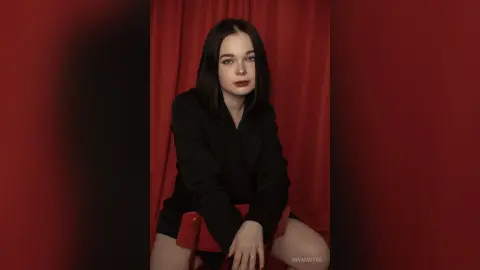 Moody portrait of a young woman in a black blazer sitting against red curtains.