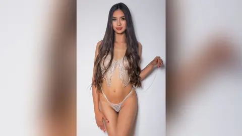 LucyMartin poses against a white wall wearing a clear, beaded string bikini that exposes her breasts and pussy.