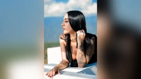 Elegant woman with long black hair in a black top posing against a scenic mountain landscape at sunset.