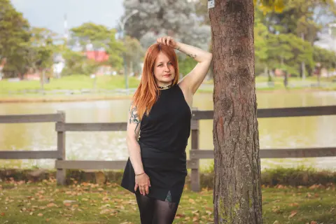 Redheaded woman in a black dress leaning against a tree by a peaceful lake in a park.